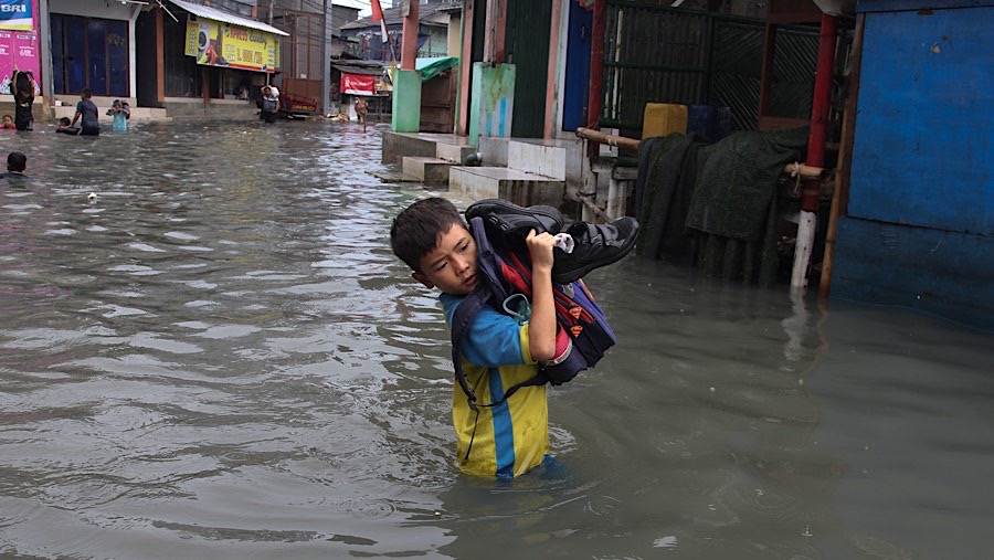 Anak-anak yang baru pulang sekolah melintasi banjir dengan menenteng tas dan sepatunya. (Bloomberg Technoz/Andrean Kristianto)