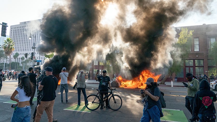 Mobil-mobil terbakar di belakang para pengunjuk rasa selama demonstrasi di Los Angeles, California, AS, Minggu (8/6/2025). (Kyle Grillot/Bloomberg)